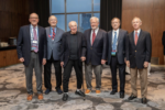 A group of six older adults, all wearing suits, standing in a room with large windows and wooden paneling. One person is using a cane. They are likely attending an event or gathering.